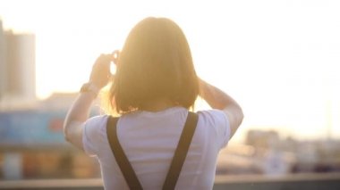 beyaz t-shirt koyu saçlı kız gün batımı arka ışık arka görünümüne karşı akıllı telefon ile büyük şehir resmi alır