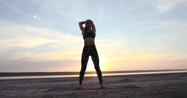 スリムな若い女性のシルエットは 美しい背中の夕日の光に対して海でヨガマットの上に立って腕の筋肉を伸ばします — ストック動画