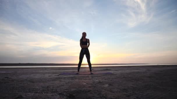 集中した女性のシルエットは 海の上に夕日に対してマットの上に立って体の筋肉を伸ばすヨガのテクニックを練習します — ストック動画