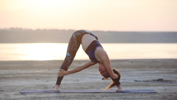 熟练的年轻女子在时髦的运动装执行先进的瑜伽姿势宽腿向前弯曲通过海上特写慢动作 — 图库视频影像