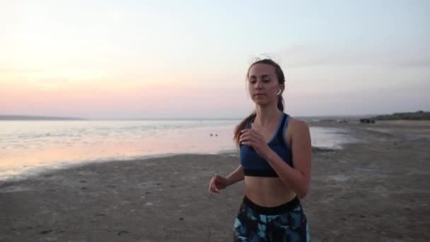 美しい夕日の空に対して海に沿ってビーチに沿ってトレンディなスポーツウェアのジョギングで集中女性は スローモーションを閉じます — ストック動画