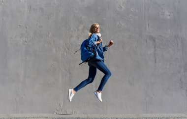 Tam vücut çanta ve cep telefonu ile yürüyüş mutlu genç kadın portresi. Gri arka planda havada atlama neşeli öğrenci