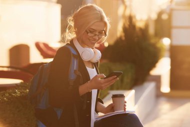 Genç yetenekli kadın öğrenci rahat giyim şehir etrafında yürüyüş giyinmiş ve kulaklıklar üzerinden radyo dinleme. Çekici esmer kadın gitmek ve mobil ile açık havada ücretsiz zaman zevk kahve almak