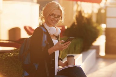 Genç yetenekli kadın öğrenci rahat giyim şehir etrafında yürüyüş giyinmiş ve kulaklıklar üzerinden radyo dinleme. Çekici esmer kadın gitmek ve mobil ile açık havada ücretsiz zaman zevk kahve almak