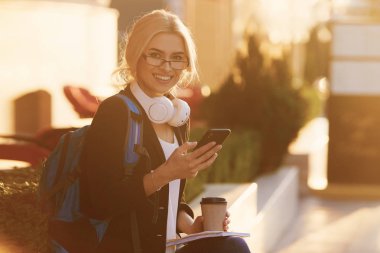 Genç yetenekli kadın öğrenci rahat giyim şehir etrafında yürüyüş giyinmiş ve kulaklıklar üzerinden radyo dinleme. Çekici esmer kadın gitmek ve mobil ile açık havada ücretsiz zaman zevk kahve almak