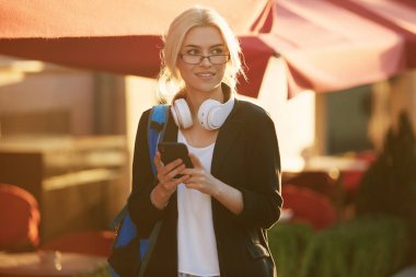 Genç yetenekli kadın öğrenci rahat giyim şehir etrafında yürüyüş giyinmiş ve kulaklıklar üzerinden radyo dinleme. Çekici esmer kadın gitmek ve mobil ile açık havada ücretsiz zaman zevk kahve almak