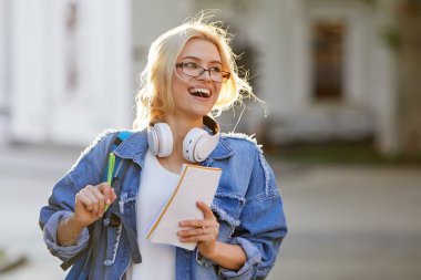 Sırt çantası ile gözlük bir gülümseyen genç kadın öğrenci portre kulaklık ve yazma notları ile müzik dinleme. Çevrimiçi ve uzaktan eğitim konsepti