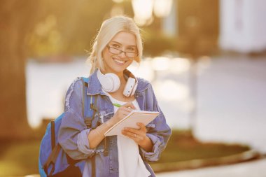 Sırt çantası ile gözlük bir gülümseyen genç kadın öğrenci portre kulaklık ve yazma notları ile müzik dinleme. Çevrimiçi ve uzaktan eğitim konsepti