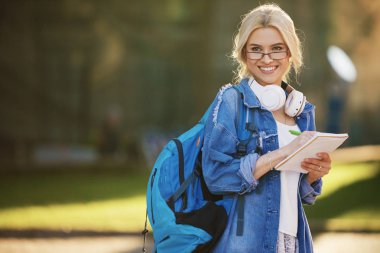 Sırt çantası ile gözlük bir gülümseyen genç kadın öğrenci portre kulaklık ve yazma notları ile müzik dinleme. Çevrimiçi ve uzaktan eğitim konsepti