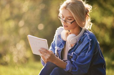 Doğa parkında oturan gözlüklü, gülümseyen bir genç kadın öğrencinin portresi. Kulaklıklarla müzik dinleme ve notlar yazma. Çevrimiçi ve uzaktan eğitim konsepti