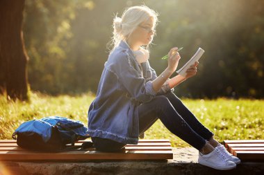 Doğa parkında oturan gözlüklü, gülümseyen bir genç kadın öğrencinin portresi. Kulaklıklarla müzik dinleme ve notlar yazma. Çevrimiçi ve uzaktan eğitim konsepti