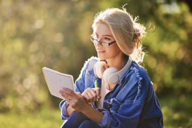 Doğa parkında oturan gözlüklü, gülümseyen bir genç kadın öğrencinin portresi. Kulaklıklarla müzik dinleme ve notlar yazma. Çevrimiçi ve uzaktan eğitim konsepti