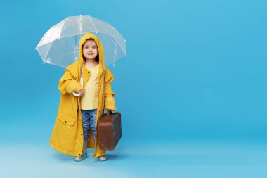 Mavi stüdyo arka planında şeffaf şemsiyesi olan mutlu komik çocuk. Kız sarı yağmurluk ve lastik çizme giyiyor. Antika bir valiz taşıyor.