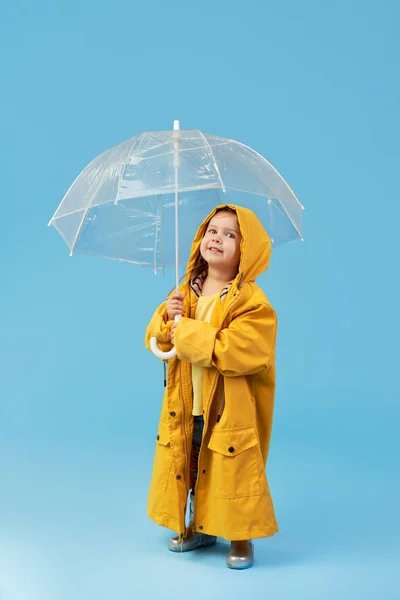 Happy funny child with transparent umbrella posing on blue studio background. Girl is wearing yellow raincoat and rubber boots. Holds a vintage travel suitcase