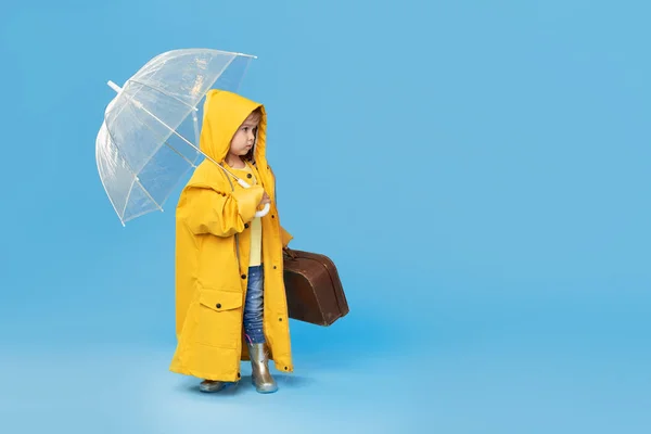 青いスタジオの背景にポーズを透明傘で幸せな面白い子 女の子は黄色のレインコートとゴムブーツを着ている ヴィンテージトラベルスーツケースを持っている — ストック写真