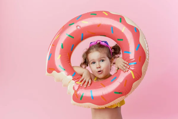 Happy surprised toddler girl in swimsuit with inflatable circle donut on a colored pink background. Wow emotions