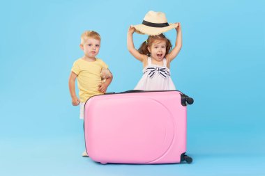 Mutlu çocuklar yaz tatili için hazırlanan renkli pembe bavul üzerinde oturuyor. Genç yolcular. Küçük kız ve çocuk, kardeş ve kardeş, mavi arka planda izole eğlenceli