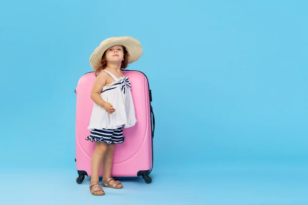 Happy child girl tourist packs clothes into a suitcase for travel, summer vacation. Toddler kid in straw hat  holding a pink suitcase on a blue background