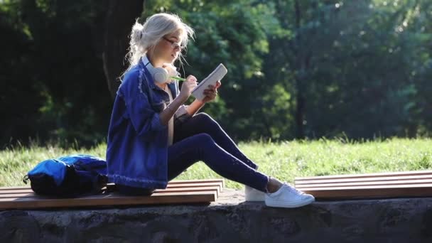 Nätt blondin sitter på brunt trä bänk skriver i anteckningsboken — Stockvideo