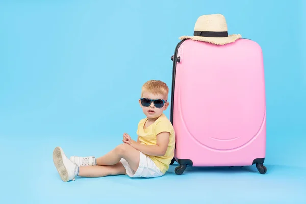 幸せな子供の男の子の観光は 夏休みのためのスーツケースに服をパックします サングラスをかけた幼児の子供は 青い背景にピンクのスーツケースの近くに座っています — ストック写真