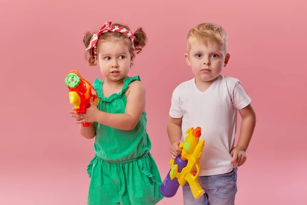 夏の服で幸せな小さな幼児の男の子と女の子は ピンクの壁の背景に隔離されたおもちゃの水銃を保持しています 子供スタジオの肖像画 人々の子供時代のライフスタイルの概念 — ストック写真