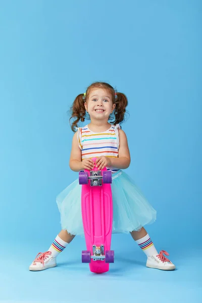 很酷的幼儿女孩在明亮的衣服举行滑板 看着相机隔离在蓝色背景 — 图库照片