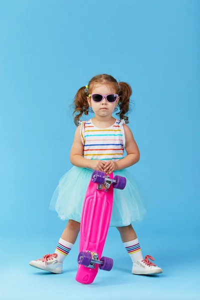 很酷的幼儿女孩在明亮的衣服举行滑板 看着相机隔离在蓝色背景 — 图库照片