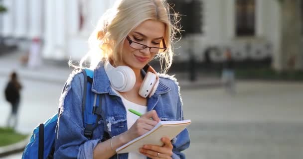 Bonito estudiante con pelo claro mira a su alrededor y escribe — Vídeos de Stock