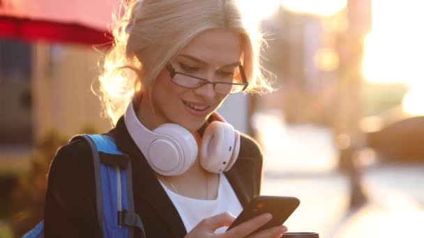 街の通りに白いヘッドフォンとスマートフォンを持つスタイリッシュな学生の女の子 — ストック動画