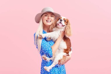 Kral Charles spaniel köpek kucaklayan yaz şapka gülümseyen genç sarışın kadın Portresi. sahibi ve evcil hayvan ilişkileri kavramı. Veteriner sağlığı. Pembe arka planda izole ön görünüm.