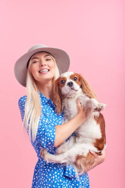 Kral Charles spaniel köpek kucaklayan yaz şapka gülümseyen genç sarışın kadın Portresi. sahibi ve evcil hayvan ilişkileri kavramı. Veteriner sağlığı. Pembe arka planda izole ön görünüm.