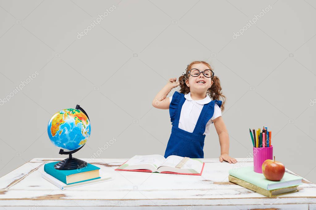 Smart toddler kid. Back to school and happy time! Cute industrious child in glasses is sitting at a desk indoors. Kid is learning in class on background of blue wall. Girl reading the book.