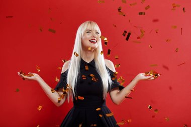 Parti zamanı! Siyah elbiseli genç kutlama yapan kadının portresi. Elinde şampanya ile büyüleyici, güzel sarışın kadın kırmızı arka plan izole. Diş telleri ve kar beyazı gülümseyen kadın