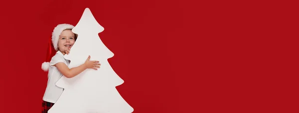 クリスマスツリーの形をした空白の看板の後ろから覗くサンタ帽子の幸せな小さな女の子 赤い背景に隔離されています テキスト用のスペース セール クリスマス 新年のコンセプト — ストック写真