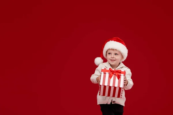 快乐的小男孩在圣诞老人红帽子拿着圣诞礼物 礼品盒 圣诞节和新年快乐 红色背景 — 图库照片