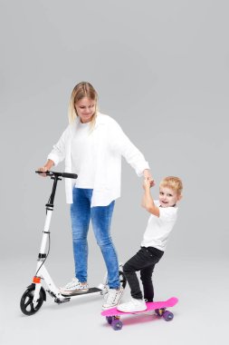 Scooter ve kaykay üzerinde duran genç kadın ve çocuk çocuk. Anne oğlu binmek öğretir, eğlenceli ve sevinçle gri arka plan üzerinde kamera bakarak var