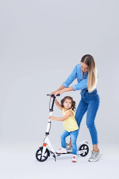 年轻女子和小女孩站在滑板车和滑板上 妈妈教女儿骑 有一个乐趣和快乐地看着相机在灰色背景 — 图库照片