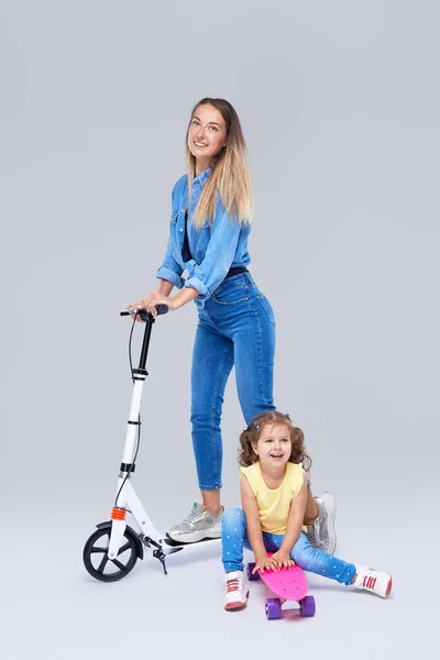 スクーターとスケートボードの上に立つ若い女性と小さな女の子 ママは グレーの背景の上にカメラで楽しさと喜びを見て 乗るために娘を教えます — ストック写真