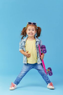 Denim giysiler cool küçük yürümeye başlayan kız mavi arka plan üzerinde izole kamera bakarak bir kaykay ve komik tutar