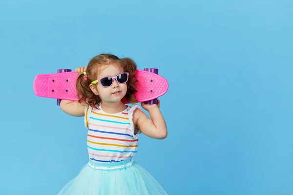 很酷的幼儿女孩在明亮的衣服和太阳镜背着滑板在他的肩膀 看着相机孤立在蓝色背景 — 图库照片