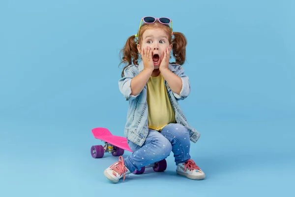 デニムの服を着たクールな幼児の女の子は スケートボードに座って 青い背景に隔離されたカメラを見て面白い — ストック写真