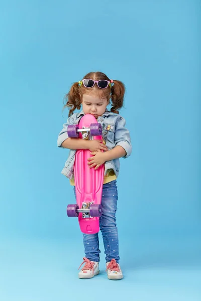 酷小蹒跚学步的女孩在牛仔服装举行滑板和有趣的看相机隔离在蓝色背景 — 图库照片