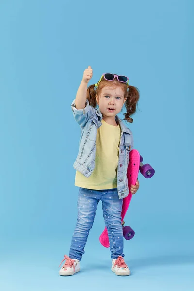 酷小蹒跚学步的女孩在牛仔服装举行滑板和有趣的看相机隔离在蓝色背景 — 图库照片