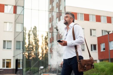 Orta yaşlı sakallı düşünen ciddi konsantre iş adamı ayakta ve elektronik sigara içiyor. Ağızdan duman üfler. Kulak içi kablosuz kulaklık. Ofis merkezi arka planı