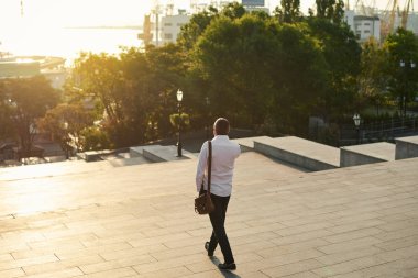 Orta yaşlı sakallı düşünen ciddi konsantre iş adamı ayakta ve elektronik sigara, vape. Ağızdan duman üfler. Kulak içi kablosuz kulaklık. Şehir arka planda gün doğumu
