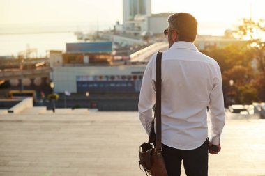 Orta yaşlı sakallı düşünen ciddi konsantre iş adamı ayakta ve elektronik sigara, vape. Ağızdan duman üfler. Kulak içi kablosuz kulaklık. Şehir arka planda gün doğumu