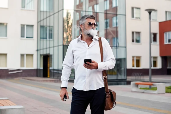 Middle-aged bearded thinking serious concentrated business man standing and smoking electronic cigarette. Blows smoke through the mouth. In-ear wireless earphone. Office centre background