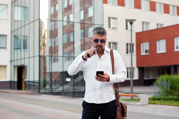 Middle-aged bearded thinking serious concentrated business man standing and smoking electronic cigarette. Blows smoke through the mouth. In-ear wireless earphone. Office centre background