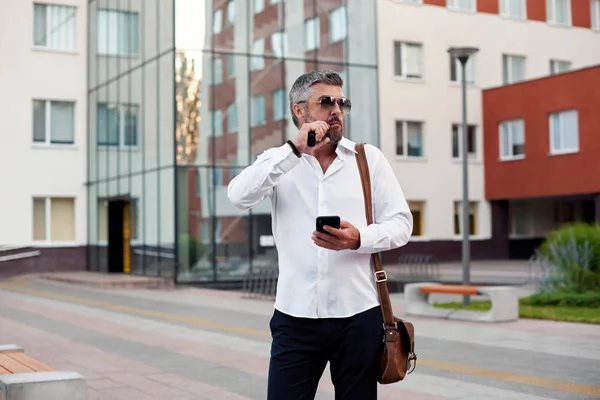 Middle-aged bearded thinking serious concentrated business man standing and smoking electronic cigarette. Blows smoke through the mouth. In-ear wireless earphone. Office centre background