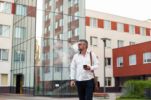 Middle-aged bearded thinking serious concentrated business man standing and smoking electronic cigarette. Blows smoke through the mouth. In-ear wireless earphone. Office centre background